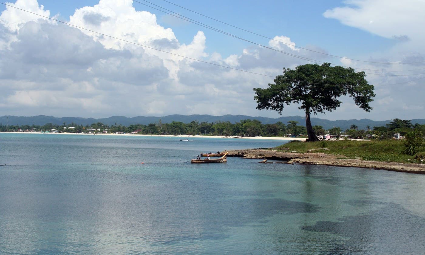 A wide, palm-fringed white sand beach stretching into the distance under a turquoise sky