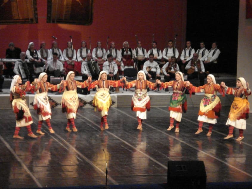 A group of dancers in colourful traditional costumes holding hands in a circle on a village square